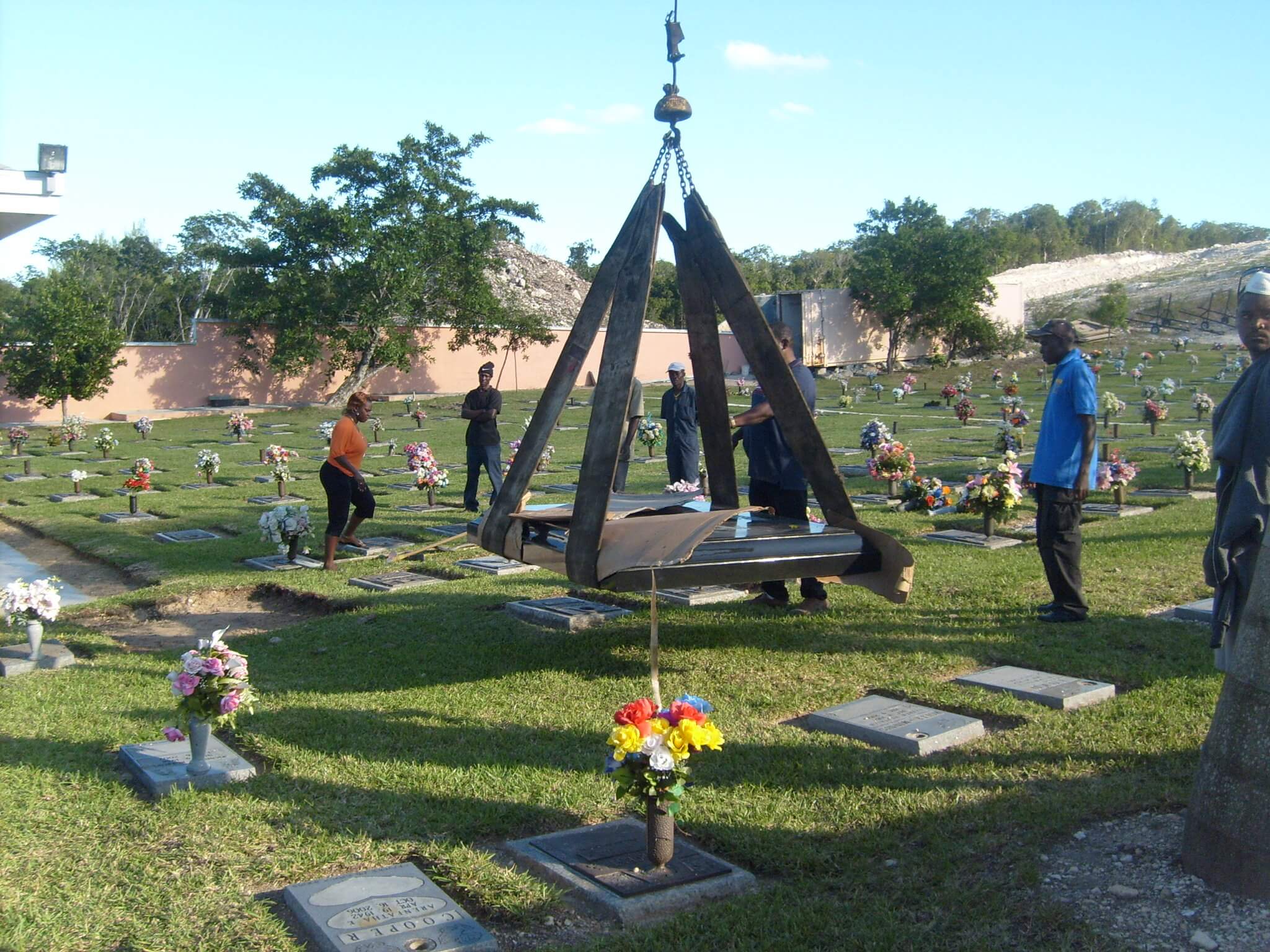 A granite monument slung beneath a crane above an open grave, with the installation crew standing by