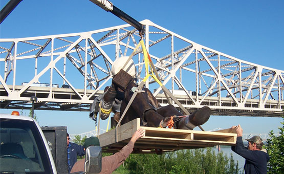 A bronze figure being lifted by crane from a truck, with a Louisville bridge over the Ohio River behind