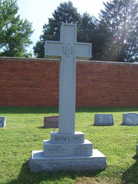Grey granite cross monument with carved IHS, standing before a brick wall