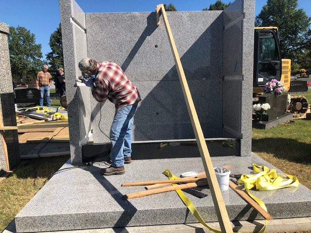 A man drilling into a granite mausoleum panel during assembly in the cemetery, with an excavator behind him